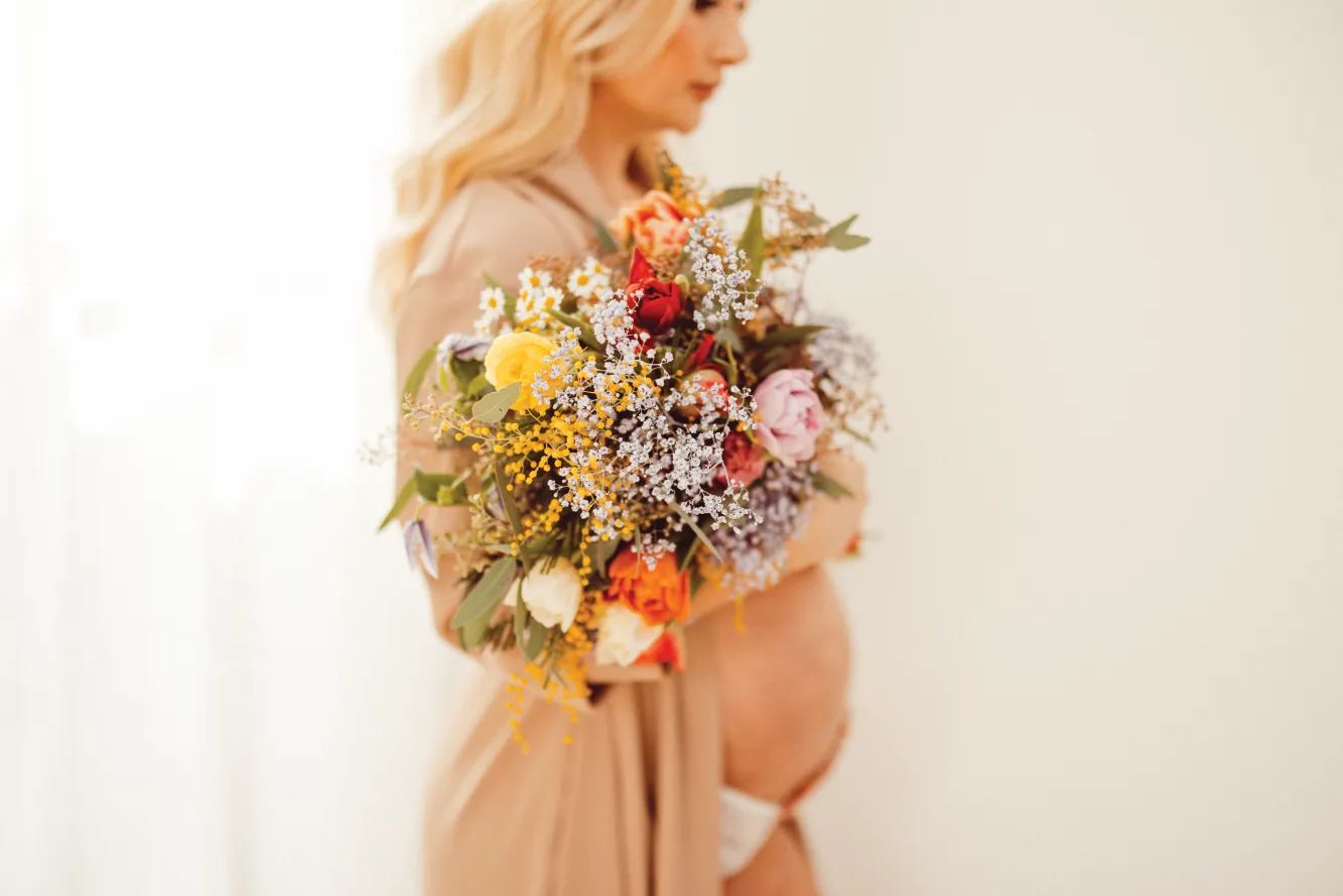 Eine schwangere Frau hält einen Blumenstrauß in ihrem Arm und berührt mit der anderen Hand ihren Babybauch.