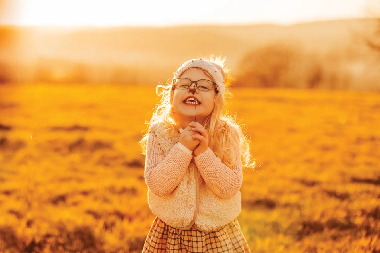 Mädchen in heller Kleidung mit Brille und einer Blume vor der Nase