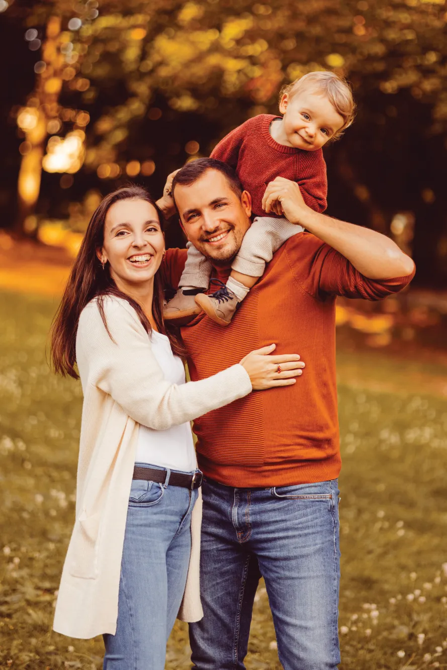 Eine dreiköpfige Familie ist in der Natur unterwegs, dabei trägt der Vater das Kind auf seinen Schultern.