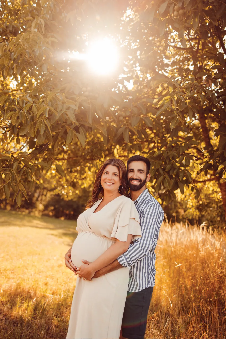 Foto in der Natur von einer Frau und ihrem Mann. Beide halten schützend ihre Hände um den Babybauch.